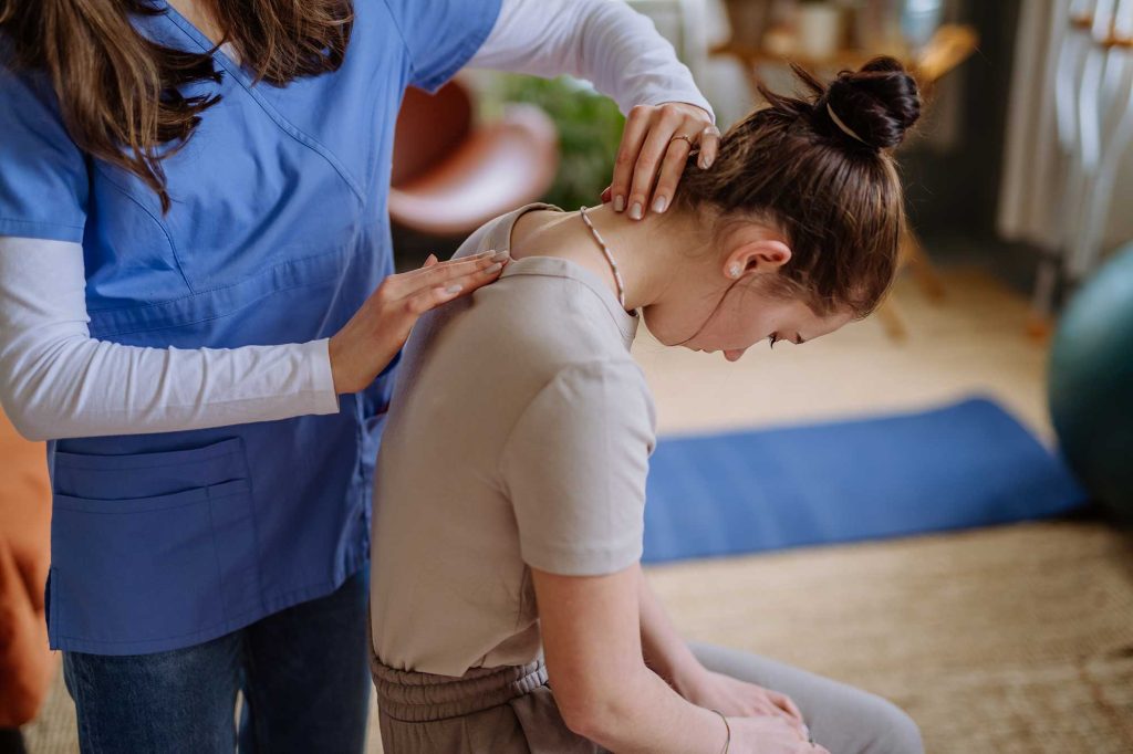 A healthcare professional examines a seated woman’s upper back and neck in a wellness clinic with a blue exercise mat nearby.