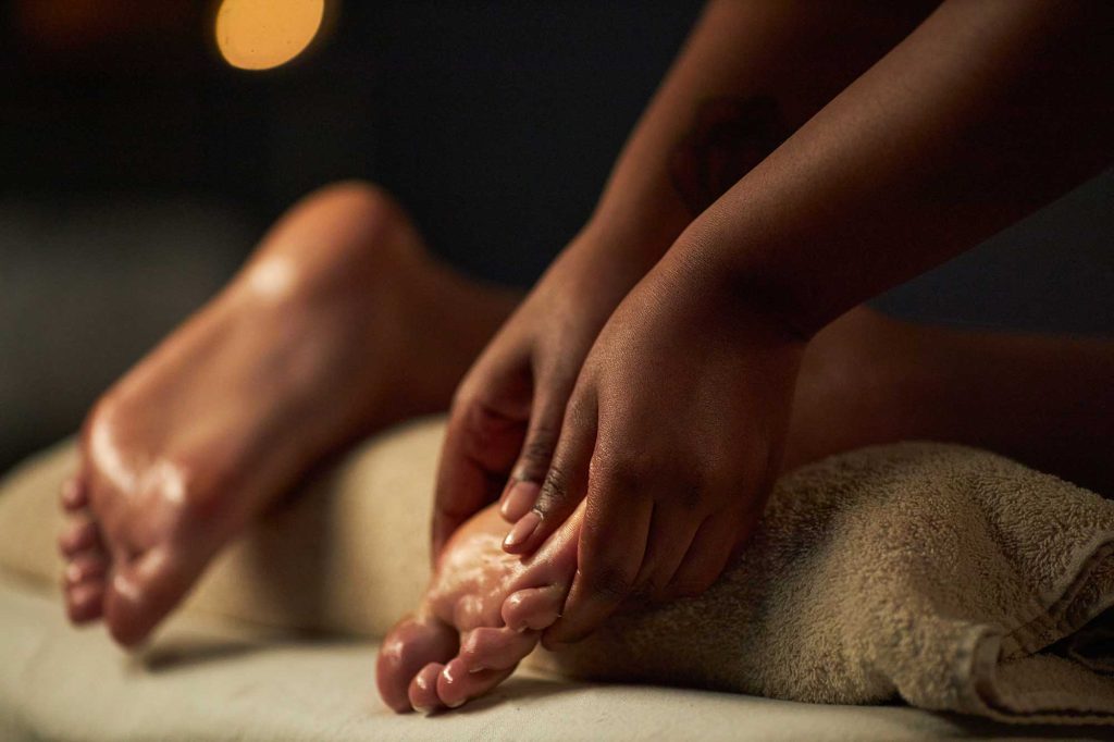 Hands massaging a person’s feet on a towel-covered spa table in warm, dim lighting.