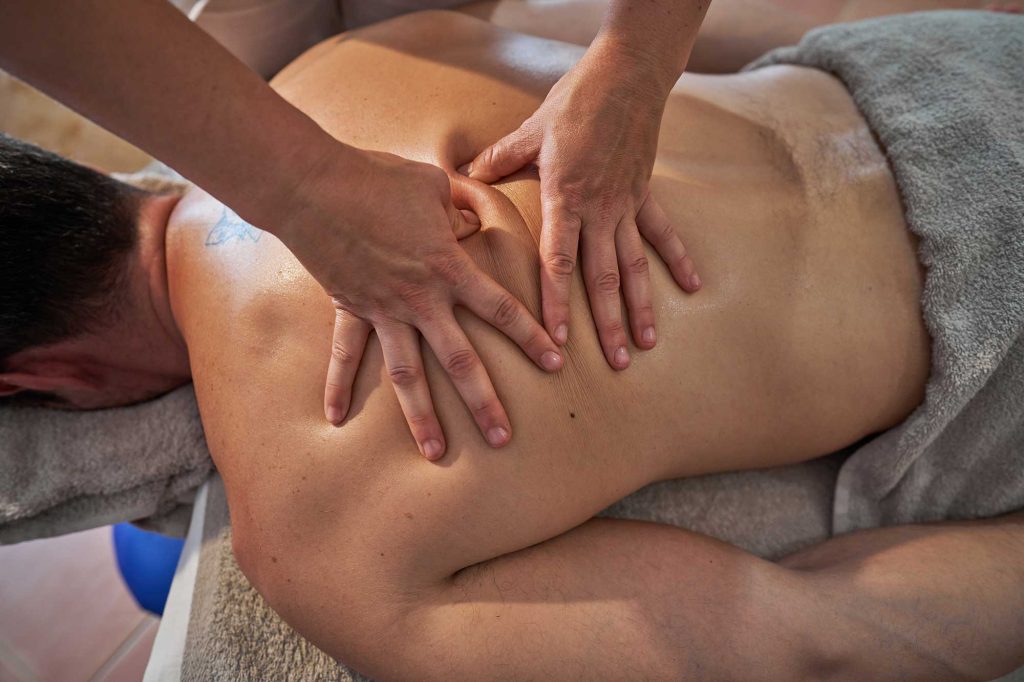 Massage therapist applying pressure to a client’s upper back and shoulder while they lie on a treatment table covered with towels.