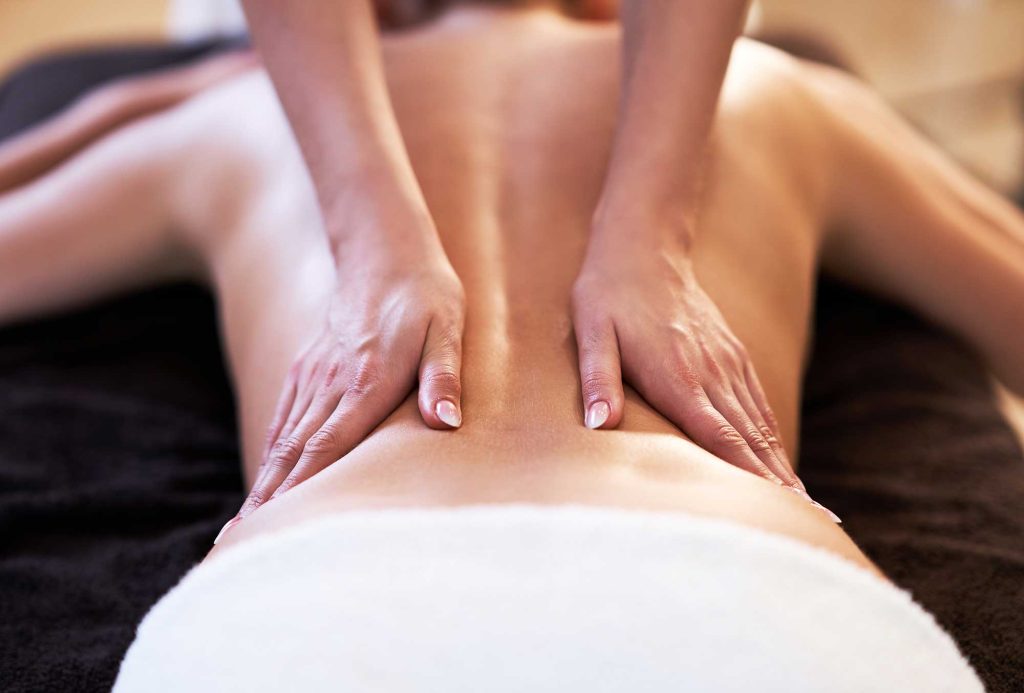 Massage therapist’s hands gently press along a client’s bare back as they lie face down under a white towel.