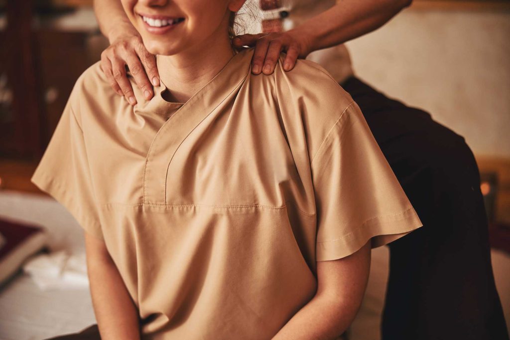 Smiling woman in tan clothing receives a shoulder massage from a person standing behind her in a warmly lit room.